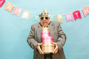 Senior man in party hat holding gift boxes against a colorful birthday banner.