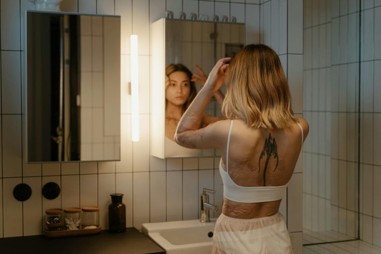A woman with tattoos and burn scars looks at her reflection in a bathroom mirror.